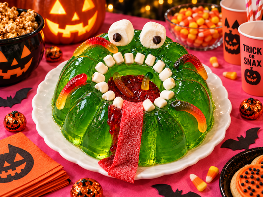Green lime gelatin monster with marshmallow eyes and teeth, gummy worms and a red candy tongue on a Halloween party table.