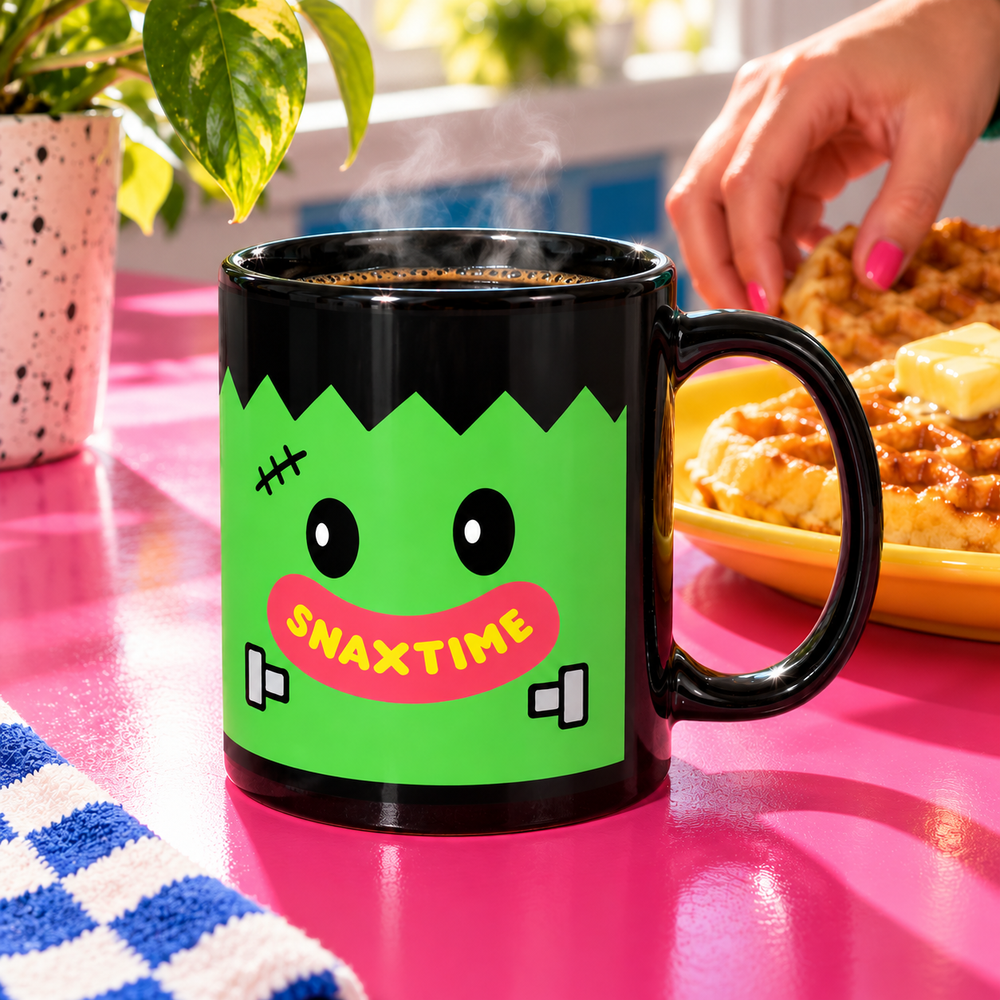 Black Snaxtime Frankenstein 11 oz mug filled with coffee on a pink breakfast counter beside waffles.